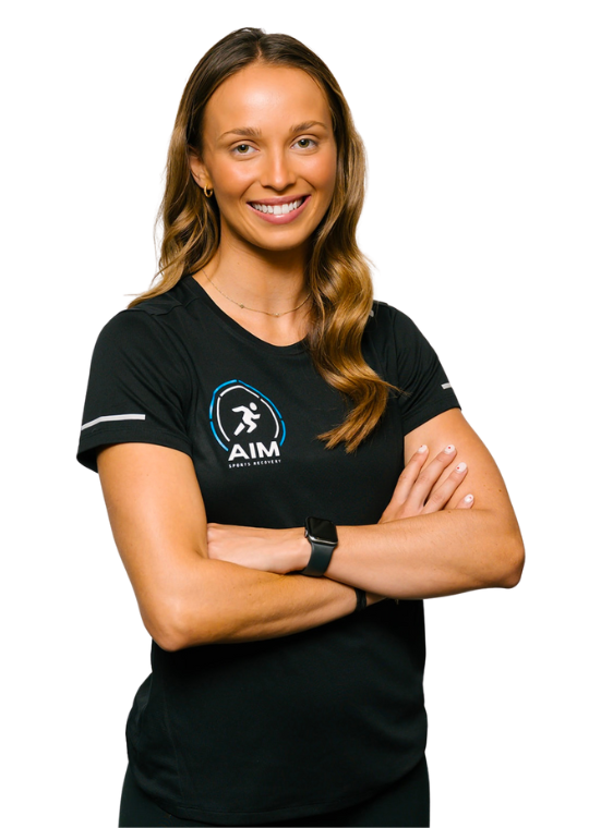 A smiling woman with long, wavy brown hair stands with arms crossed. She wears a black AIM Sports Center t-shirt and a smartwatch.