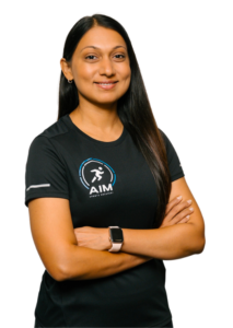 A woman with long dark hair stands with arms crossed, smiling. She wears a black AIM Sports Medicine t-shirt and a smartwatch, posed against a plain background.