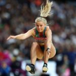 A female athlete with long blonde hair is mid-air during a long jump in a stadium. She is wearing a green and yellow uniform with a visible number. Her arms are forward, legs bent, and audience members are blurred in the background.