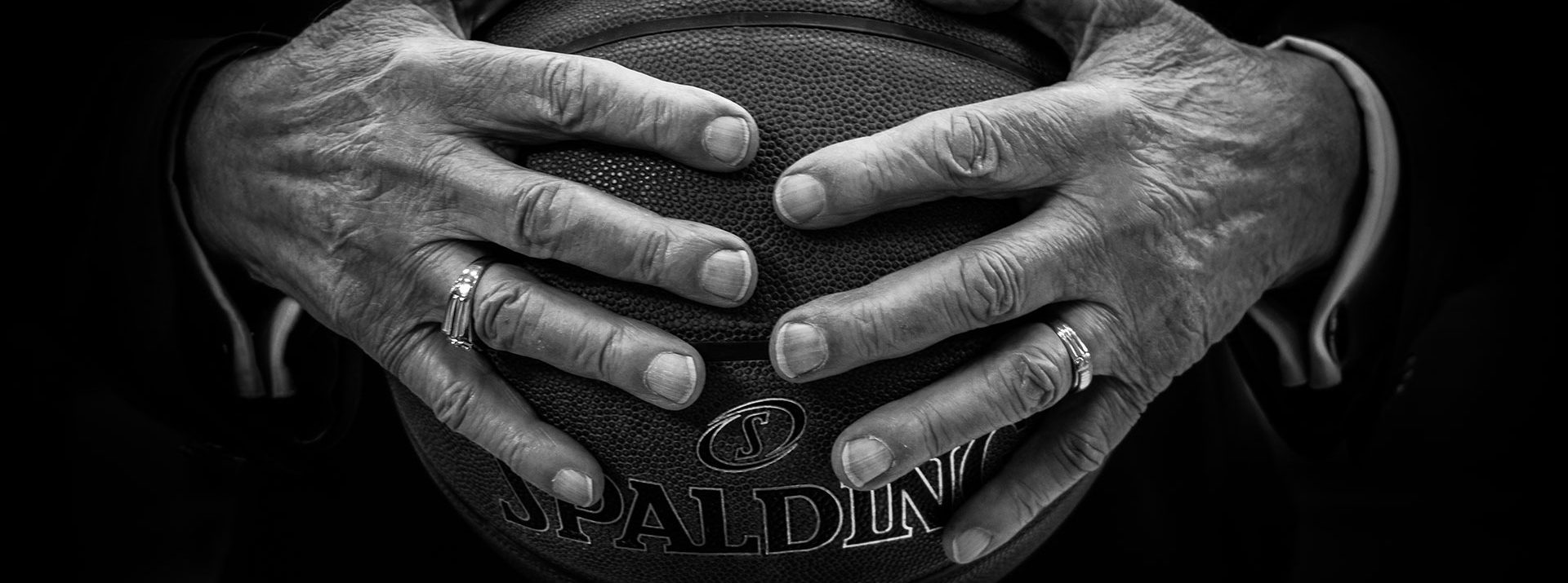 A black and white photo showing a close-up of a person’s hands holding a basketball. The hands, adorned with rings, are pressing firmly into the Spalding basketball, creating a sense of strength and determination pivotal in health & sports recovery. The background is dark, highlighting the hands and ball.