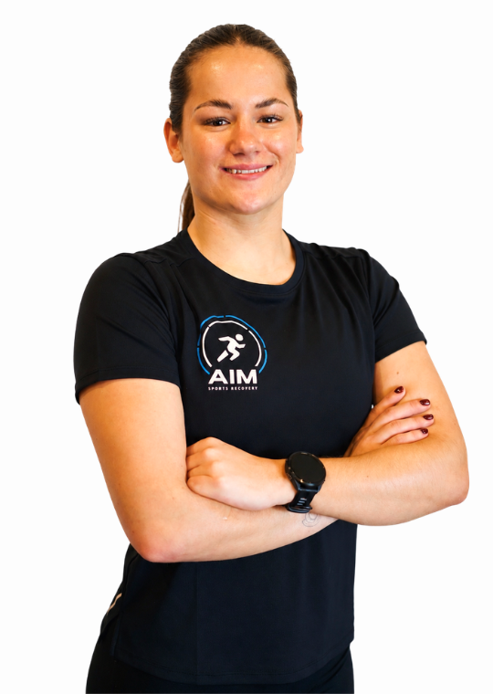 A woman with brown hair in a ponytail, wearing a black AIM Sports Science shirt and a watch, stands smiling with arms crossed against a white background.
