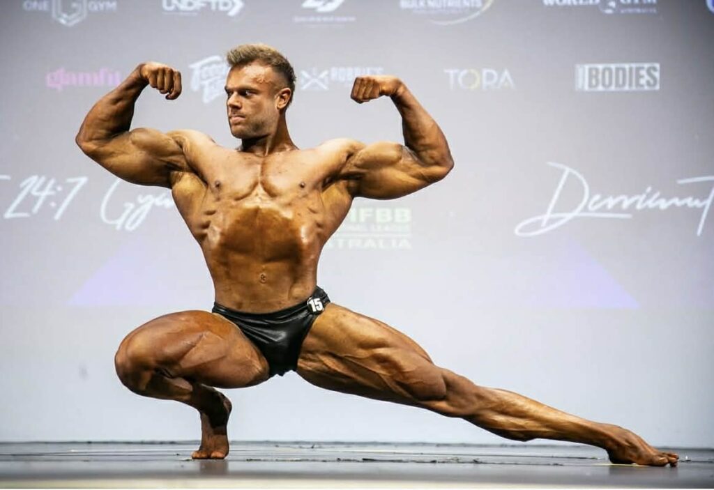 A male bodybuilder in dark posing trunks strikes a flexed side lunge pose on stage, showcasing muscular definition under bright lights, with a blurred competition backdrop—an impressive scene for potential athlete sponsorship.
