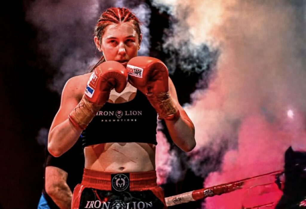 A female boxer with braided hair, wearing red gloves and black athletic gear, stands in a boxing ring with smoke and dramatic lighting, looking focused and ready to fight—an inspiring scene perfect for athlete sponsorship campaigns.