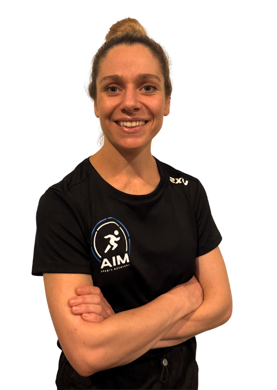 A woman with light brown hair tied up, wearing a black AIM t-shirt, stands with arms crossed and smiles at the camera against a white background.