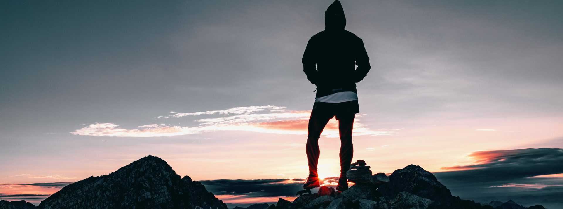 Silhouetted figure in a hoodie stands on a rocky mountain peak, gazing at a dramatic sunset with vibrant clouds and a distant horizon, creating a serene and contemplative mood perfect for health & sports recovery.