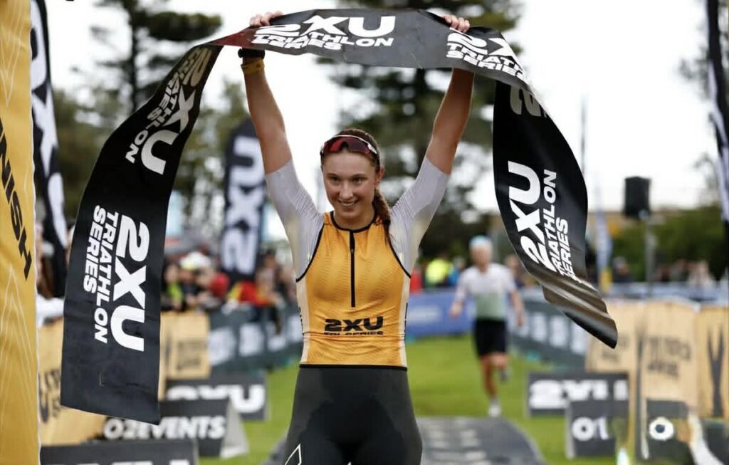 A female athlete in a yellow and black suit smiles as she raises a "2XU Triathlon Series" banner above her head, celebrating her victory at the race finish line, showcasing the impact of athlete sponsorship. Other competitors and banners are visible in the background.