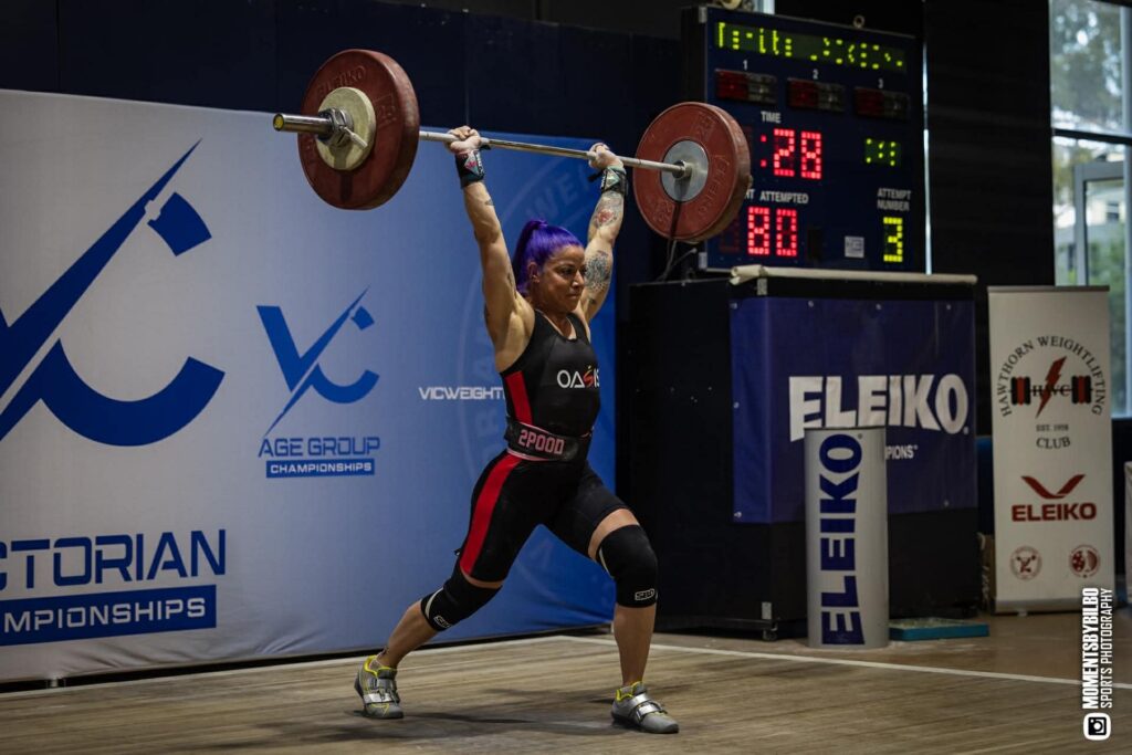 A female weightlifter with purple hair lifts a barbell overhead on a competition platform, wearing a black and red uniform. Digital scoreboards and Athlete Sponsorship banners are visible in the background.